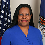 Charniele Herring standing in front of the United States flag on her right and the Virginia State flag on her left while smiling and wearing a blue blazer.